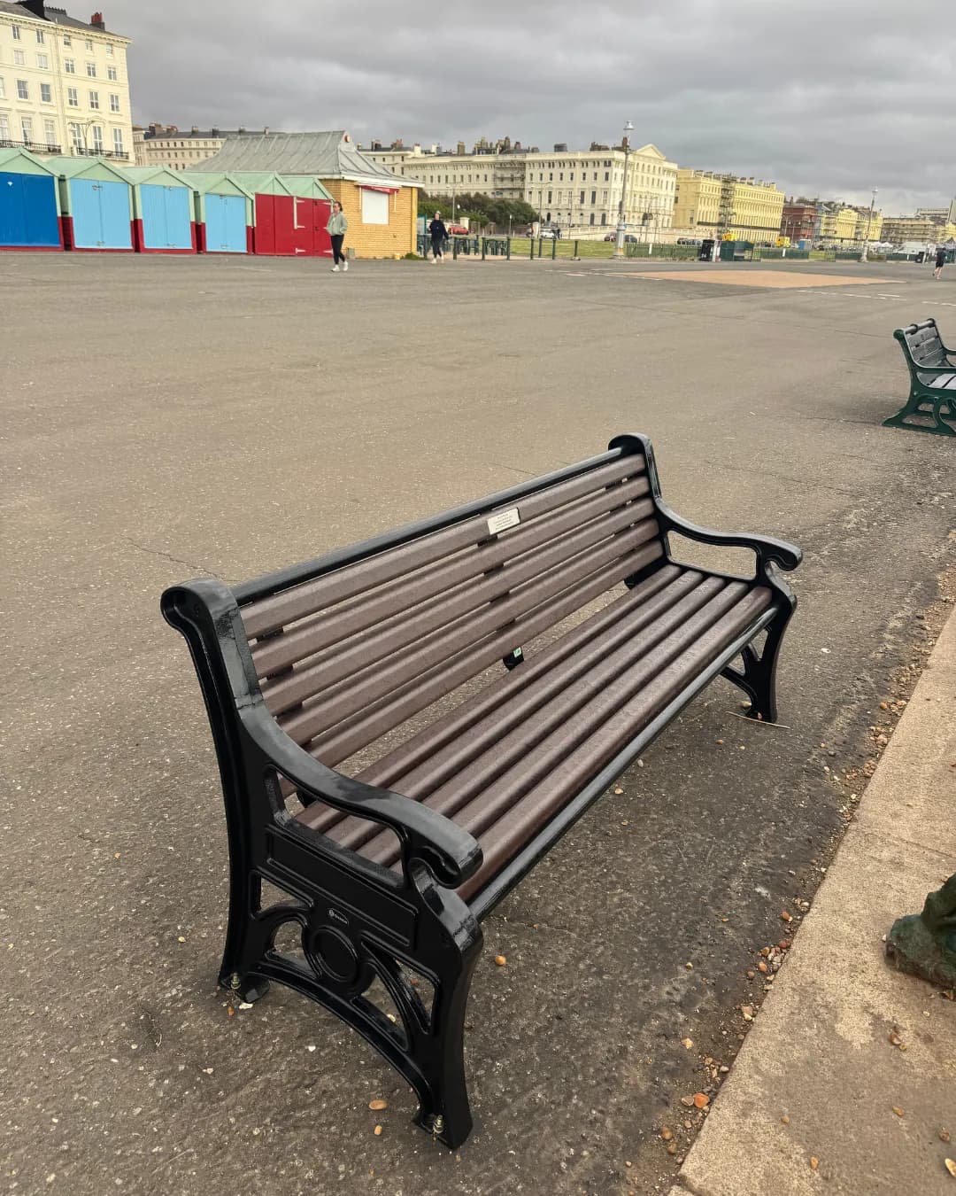 A wooden and cast iron park bench sits empty on a paved promenade with colorful beach huts and buildings in the background under an overcast sky.