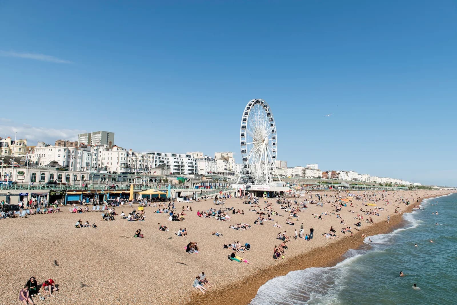 A wide view of a crowded beach with a Ferris wheel and buildings in the background under a clear blue sky.
